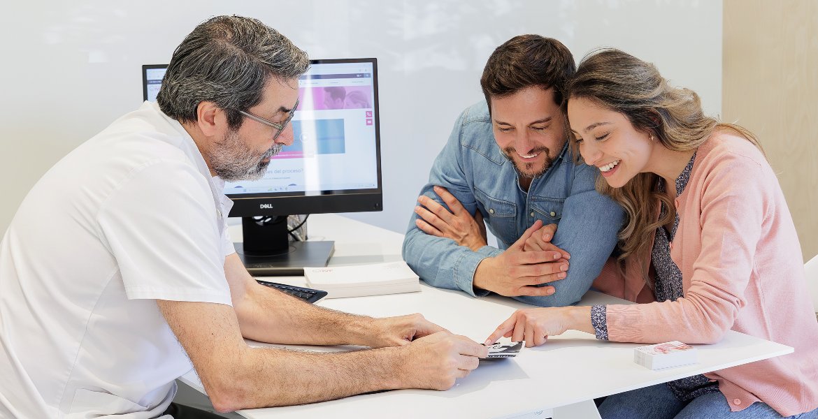 A doctor sits at a desk with a couple, discussing information and showing them documents in a modern medical office. A computer monitor and paperwork are visible on the desk.