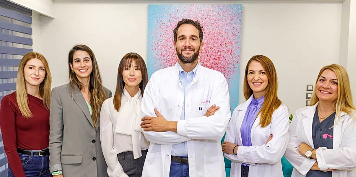 Group photo of fertility specialist Panagiotis Drakopoulos standing with his reproductive medicine team in a modern clinic interior, wearing medical uniforms and smiling at the camera