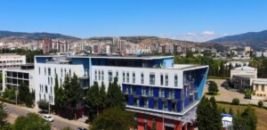 Exterior view of the LeaderMed Clinic, a modern multi-story medical facility with a blue and white facade, featuring a rooftop terrace and set against a city and mountain background.